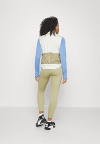 Könnyű, krém színű mellény bézs részletekkel, kék hosszú ujjú toppal és zöld leggings-szel párosítva. Sportos, laza dizájn jellemzi.