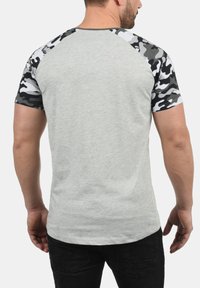 Grå T-skjorte med svart og hvit camo-mønster på ermene, kort raglan-kutt, blanding av bomullsmateriale, jevn tekstur, ingen synlige logoer.