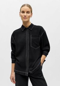 Femme aux cheveux blonds portant une chemise noire ample avec des coutures contrastées blanches et un pantalon noir assorti, debout avec une main dans la poche.