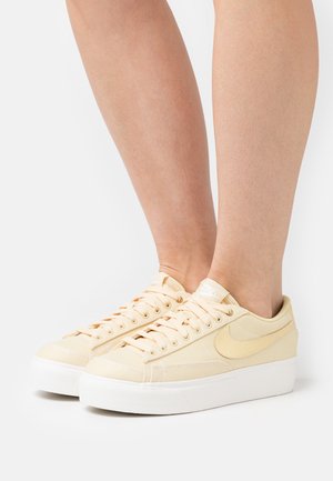 Zapatillas de lona amarillo claro con suelas blancas, que presentan un diseño clásico con cordones, acentos cosidos y un logo sutil en la lengüeta.