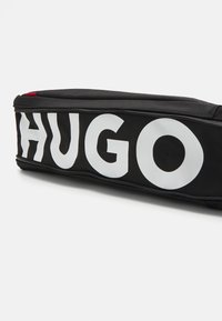 Sac en tissu noir avec une texture lisse et le logo "HUGO" en lettres blanches audacieuses. Il dispose d'une fermeture éclair et d'un petit accent rouge sur le côté.
