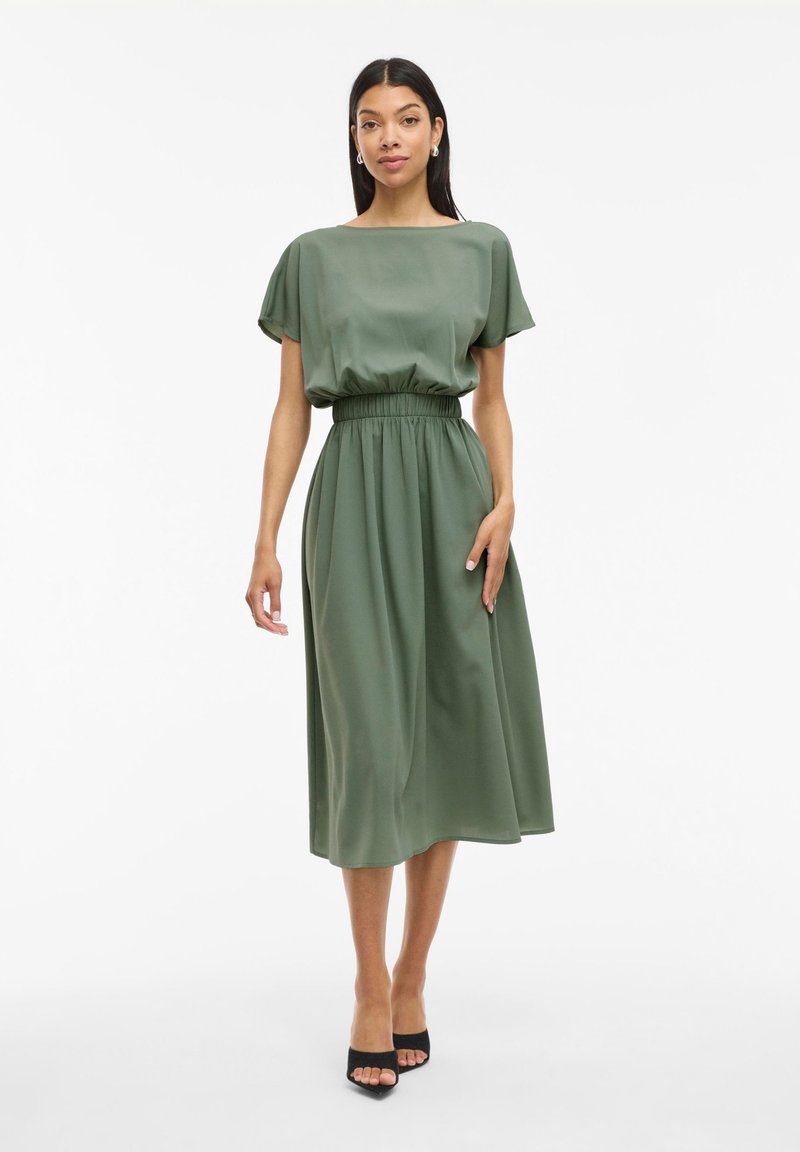Vestido midi verde de tela suave con escote barco, mangas cortas y cinturilla elástica, combinado con tacones negros.