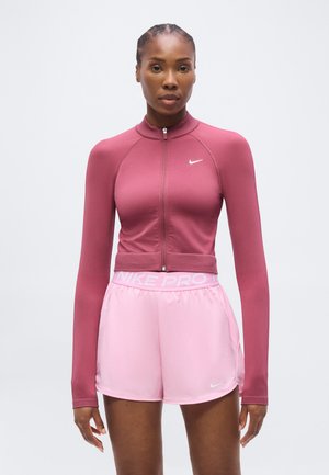 Žena v dlhom rukáve, bordo športovom tričku so zipsom a svetloružových šortkách Nike Pro na jednoliatom bielom pozadí.