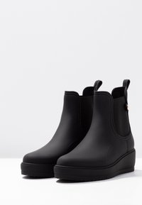 Botas de tobillo de goma negra con paneles laterales elásticos y tiradores. Presentan una suela de cuña y una banda de rodadura texturizada para mayor agarre. Acabado mate suave.