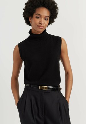 Vrouw met kort krullend haar, gekleed in een mouwloze zwarte coltrui en zwarte broek met riem, staand met handen in de zakken.