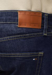 Mørkeblå denimjeans med gul søm, en liten flagglogo på lommen og en brun lærpatch merket "TOMMY JEANS" på midjen.