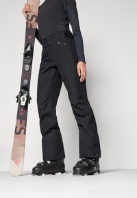 Schwarze Skihosen mit taillierter Passform, kombiniert mit schwarzen Ski-Stiefeln und einem Snowboard. Merkmale sind ein elegantes Design und verstärkte Unterschenkel.