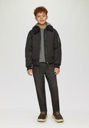 Jongen met krullend haar, gekleed in een zwarte jas met schapenvachtkraag, een groene hoodie, donkere spijkerbroek en een paar bruine sneakers, staande tegen een effen achtergrond.