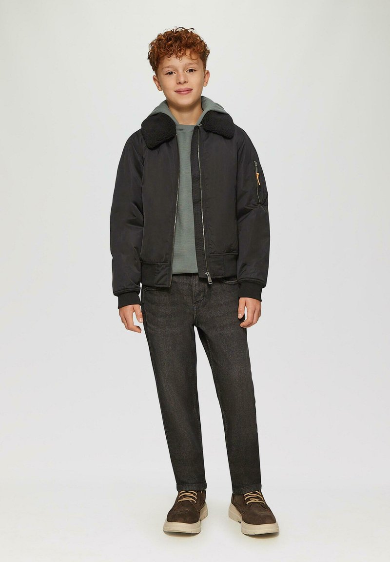 Ragazzo con capelli ricci che indossa una giacca nera con colletto in shearling, felpa verde, jeans scuri e sneakers marroni, in piedi contro uno sfondo semplice.