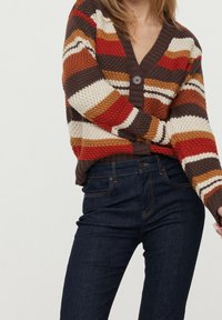 Striped cardigan in earthy tones of red, brown, and cream, featuring a v-neck, knitted texture, and single-button closure. Worn over dark jeans.