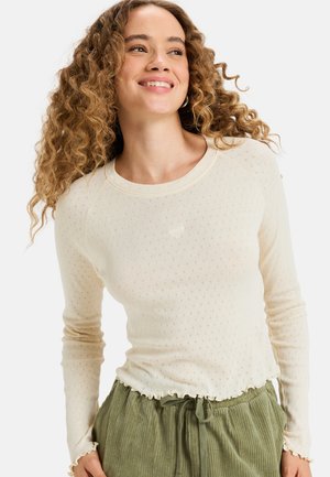 Jeune femme aux cheveux bouclés sourit, vêtue d'un haut à manches longues texturé crème et d'un pantalon en velours côtelé vert avec cordon de serrage, sur fond blanc.