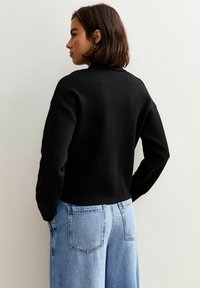 Czarny dzianinowy sweter z wysokim dekoltem i opadającymi ramionami, w połączeniu z jasnoniebieskimi spodniami o szerokich nogawkach, które mają klasyczny pięciopocketowy krój.