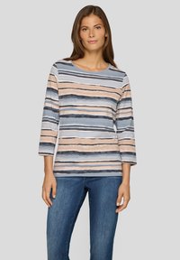 Frau trägt ein langärmliges gestreiftes Shirt in Blau-, Beige- und Schwarztönen, kombiniert mit blauen Jeans, vor einem schlichten grauen Hintergrund stehend.