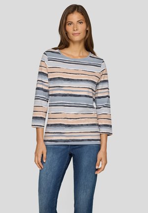 Frau trägt ein langärmliges gestreiftes Shirt in Blau-, Beige- und Schwarztönen, kombiniert mit blauen Jeans, vor einem schlichten grauen Hintergrund stehend.