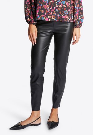 Frau trägt schwarze schmale Hosen aus Kunstleder, langärmeliges florales Oberteil und schwarze spitze Slingback-Flats vor weißem Hintergrund.