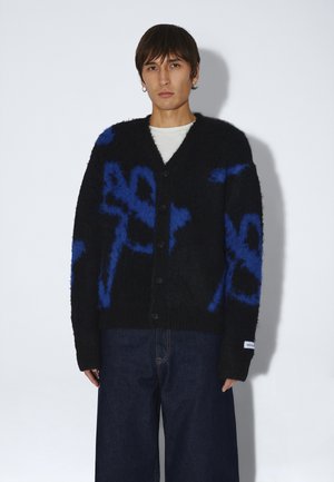 Persona con capelli corti e scuri indossa un cardigan nero abbottonato con motivi astratti blu e jeans scuri a gamba larga su sfondo bianco.