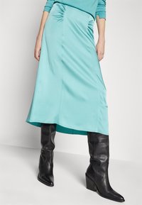 Une jupe turquoise, arrivant au genou, avec des détails froncés sur le côté, associée à de hautes bottes en cuir noires à talons. Tissu lisse et extensible.