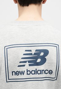 Άτομο που φαίνεται από πίσω, φοράει ανοιχτό γκρι φούτερ με σκούρο μπλε λογότυπο New Balance και κείμενο στο πάνω μέρος της πλάτης.