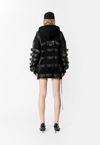 Sudadera negra con mangas oversized que presenta detalles de cinta negra, capucha con cordones y una pequeña etiqueta en la parte posterior. Se ven pantalones cortos y zapatos de tacón.