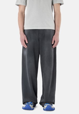 Person trägt lockere dunkelgraue Hose und blau-graue klobige Sneakers, steht mit entspannt an den Seiten hängenden Armen vor einem schlichten Hintergrund.