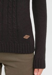 Pull en maille torsadée marron foncé avec des poignets et un ourlet côtelés. Présente une surface texturée et un petit patch en cuir avec logo en bas à gauche.