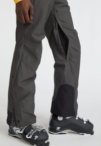 Pantalones de esquí grises con ventilaciones con cremallera, parches de rodilla reforzados en negro y un diseño ajustado, usados con botas de esquí negras que presentan herrajes plateados.