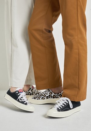 Twee mensen dragen casual sneakers—een paar zwart-wit, het andere paar met luipaardprint—staan dicht bij elkaar, gekleed in bruine en witte broeken.