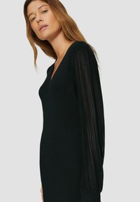 Robe noire avec un décolleté en V, corps ajusté et longues manches transparentes plissées. Texture lisse et éclat subtil sur le tissu.