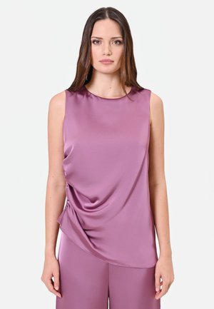 Donna con lunghi capelli castani che indossa un top senza maniche in satin color malva e pantaloni abbinati, in piedi davanti a uno sfondo chiaro e semplice.