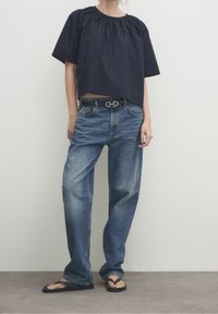 Top corto navy con scollatura arricciata, abbinato a jeans a gamba larga di colore azzurro chiaro. Footwear presenta sandali neri. Design semplice e casual.