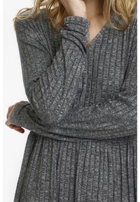 Vrouw in een donkergrijze gebreide top met lange mouwen en textuur met verticale strepen, met gekruiste armen en een hand die haar gezicht aanraakt.