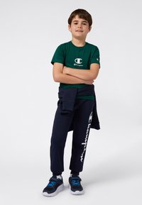 Ragazzo in piedi con le braccia incrociate, indossa una maglietta verde Champion, pantaloni tuta blu scuro con logo, scarpe coordinate e una felpa annodata in vita.
