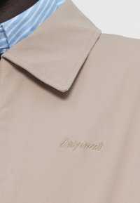 Μπεζ εξωτερικά ρούχα με υφή, που διαθέτουν ευρύ γιακά. Φαίνεται ανοιχτό μπλε πουκάμισο με ρίγες και κεντημένο λογότυπο.