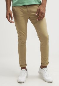 Khaki slim-fit chino kalhoty s hladkým materiálem, srolované nohavice, v kombinaci s bílými texturovanými teniskami a zelenou košilí.