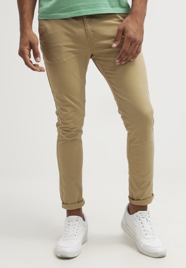 Khaki slim-fit chino kalhoty s hladkým materiálem, srolované nohavice, v kombinaci s bílými texturovanými teniskami a zelenou košilí.