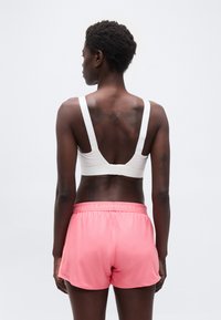 Γυναίκα με κοντά μαλλιά, φορώντας λευκό αθλητικό σουτιέν και ροζ αθλητικά σορτς Under Armour, εμφανίζεται από πίσω με φόντο ένα απλό φόντο.