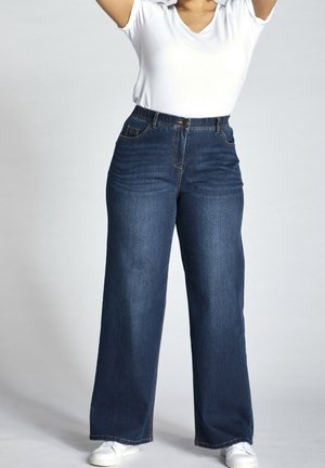 Person iført højtaljede, vidde mørkeblå jeans med en hvid t-shirt og hvide sneakers, med armene hævet over hovedet.