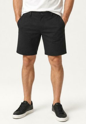 Man die zwarte casual shorts en zwarte sneakers met witte zolen draagt, staat met zijn handen in zijn zakken tegen een effen achtergrond.