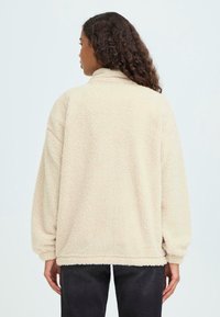 Beež villane poolkampsun kõrge kaeluse, langenud �õlgade ja soonikute varrukatega. Pehme tekstuur ja lõdva istuvusega, nähtud tagantpoolt.