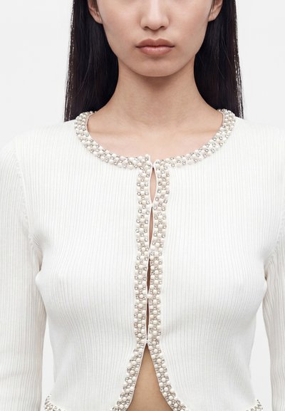 Femme portant un cardigan blanc côtelé avec bordure de perles et de strass le long de l'encolure et de l'ouverture frontale, le devant est en grande partie déboutonné.