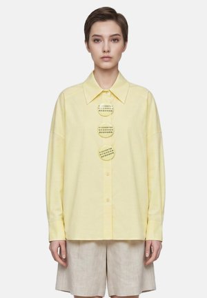 Vrouw draagt een lichtgele blouse met knopen met drie grote ronde knopen met cijfers aan de voorkant, gecombineerd met beige korte broek.