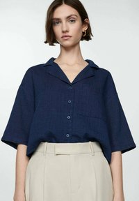 Navy blue short-sleeve button-up shirt with a collar, featuring a front pocket and textured fabric. Paired with light beige high-waisted trousers.