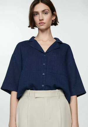 Navy blue short-sleeve button-up shirt with a collar, featuring a front pocket and textured fabric. Paired with light beige high-waisted trousers.