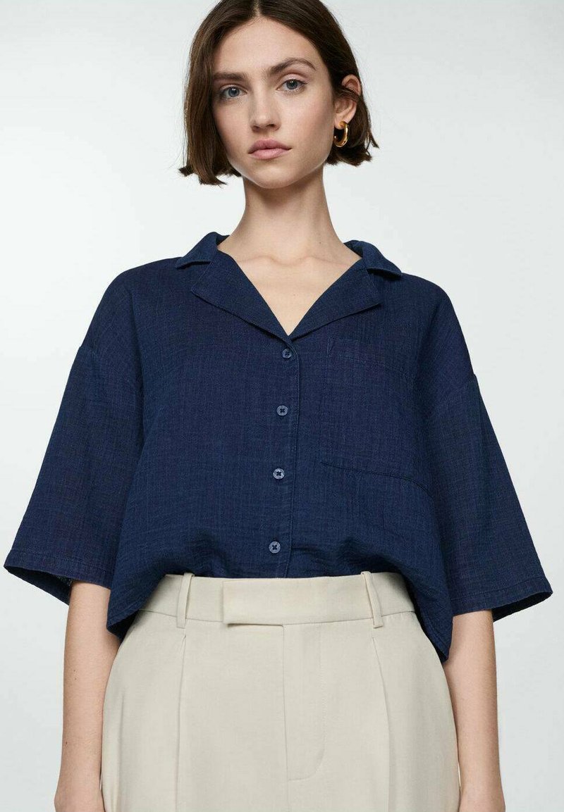 Navy blue short-sleeve button-up shirt with a collar, featuring a front pocket and textured fabric. Paired with light beige high-waisted trousers.