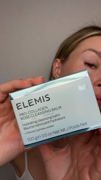 Henkilö pitää käsissään Elemis Pro-Collagen Rose Cleansing Balm -purkkia, jossa näkyvät tuotedetaljit ja paino 100 grammaa.