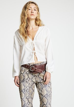 Blouse blanche à manches longues avec cordon de serrage à l'avant et bordure en dentelle, associée à un pantalon ajusté à imprimé serpent et une ceinture marron à clous.