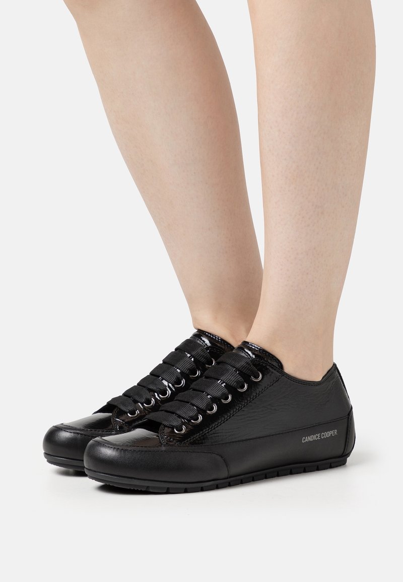 Jambes portant des baskets basses en cuir noir avec des lacets larges et le logo "Candice Cooper" sur le côté sur fond blanc.