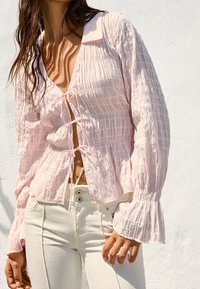 Woman wearing a light pink textured blouse with ties down the front and cream high-waisted pants, standing against a white wall.