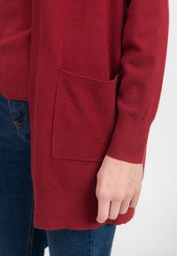 Rød strikket cardigan med en tekstureret overflade, åben front og sidelommer, parret med denimbukser. Ærmet har en ribbet manchet.