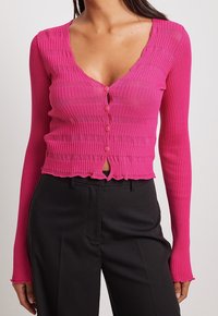 Eng anliegender, gerippter, rosa Cardigan mit langen Ärmeln, V-Ausschnitt, Knopfverschluss und einem gewellten Saum. Kombiniert mit einer schwarzen Hose.